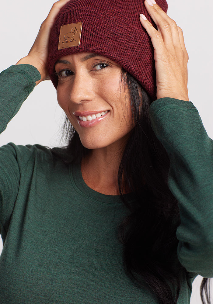 A person wearing a green long-sleeve shirt, dark hair flowing, smiles while holding the sides of a Cranberry Melange Baylor Beanie by Woolx. The beanie, crafted from merino wool, displays a simple leather patch design.