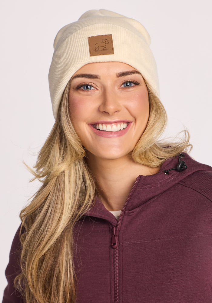 A smiling woman with long blonde hair wears the Woolx Baylor Beanie in Sweet Cream, featuring a brown patch, paired with a maroon zip-up jacket. The background is plain and light-colored.