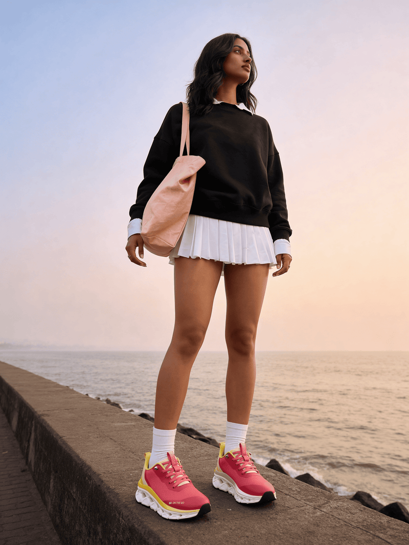 Woman standing by the ocean wearing a black sweater, white skirt, bacca bucci sneakers, and a beige backpack.