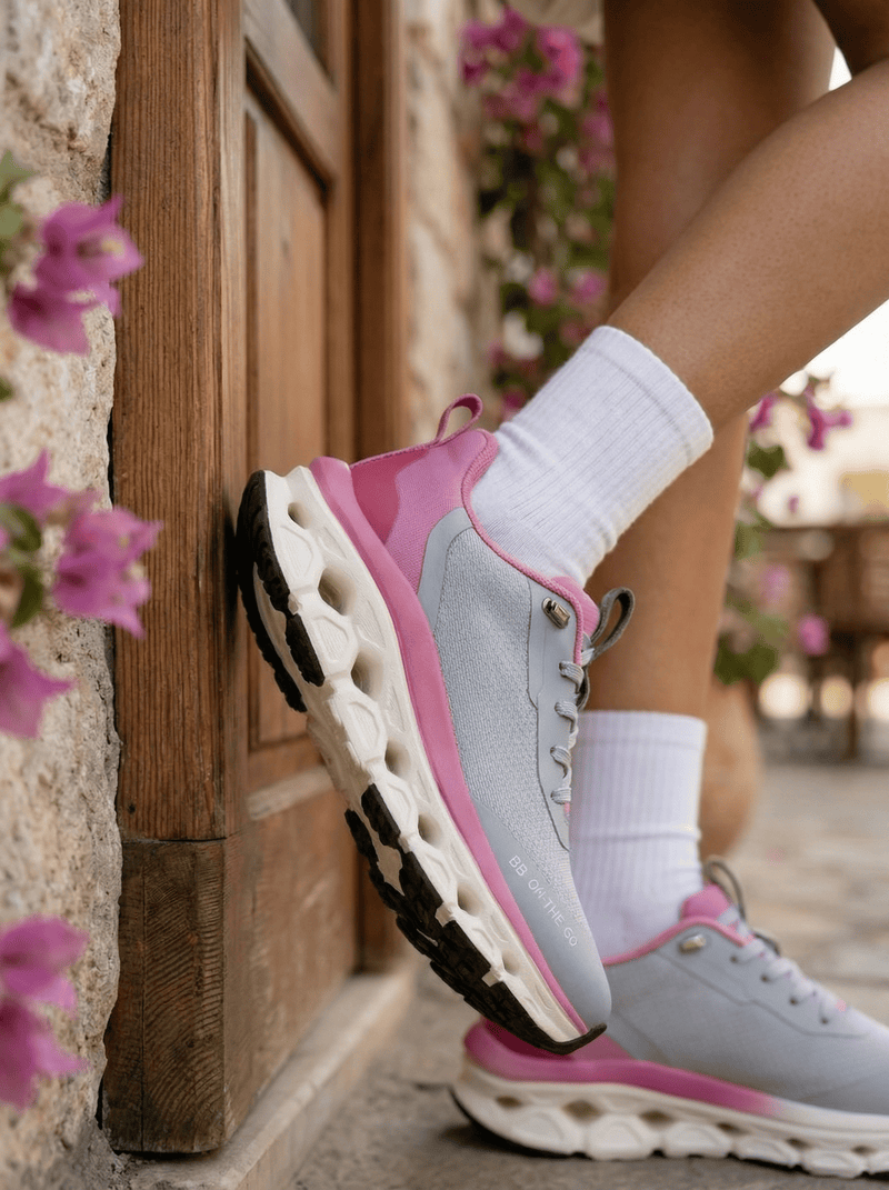 Person wearing bacca bucci pink and gray sneakers with white socks, standing against a stone wall with flowers.