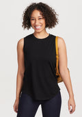 A person with curly hair smiles while wearing the sleeveless Bella Tunic Top in black by Woolx, paired with dark jeans. They carry a tan shoulder bag against a plain white background. | Tori is 5'7", wearing a size S