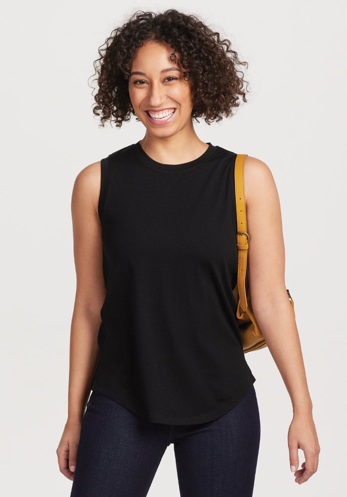 A person with curly hair smiles while wearing the sleeveless Bella Tunic Top in black by Woolx, paired with dark jeans. They carry a tan shoulder bag against a plain white background. | Tori is 5'7", wearing a size S