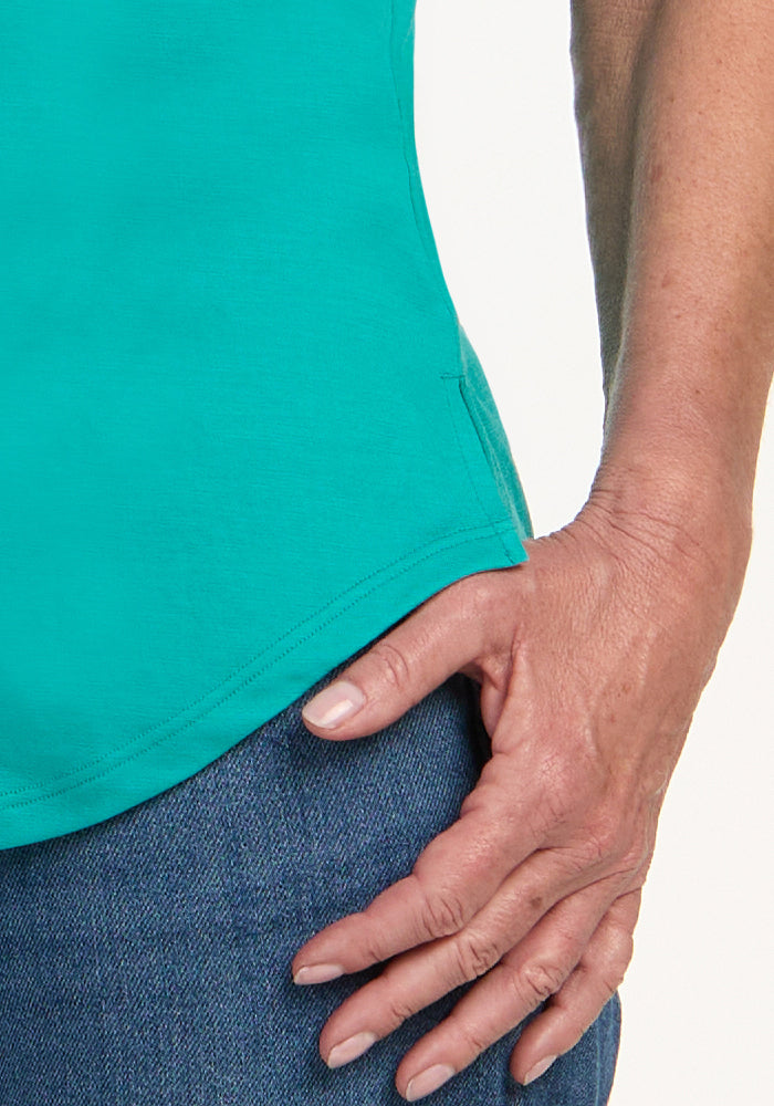 A person’s hand rests on their thigh, wearing the Woolx Bella Tunic Top in Cool Jade and blue jeans.