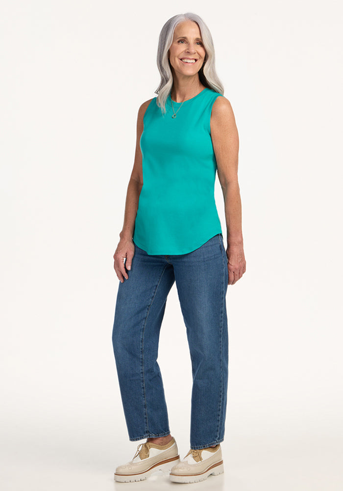 Smiling older woman wears Woolx Bella Tunic Top in Cool Jade, blue jeans, and beige shoes, standing on white background.