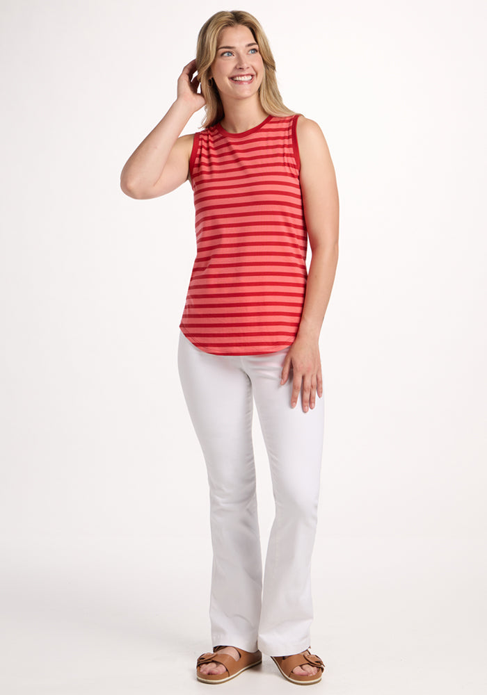 A woman with light brown hair smiles, touching her head as she stands against a plain background wearing the Woolx Bella Tunic Top in Wildberry Stripe, paired with white pants and brown sandals.