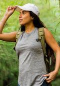 A woman wearing the Woolx Bella Tunic Top in Graphite Heather, a white cap, and a backpack stands outdoors in a lush green forest, looking to the side while holding the brim of her cap.