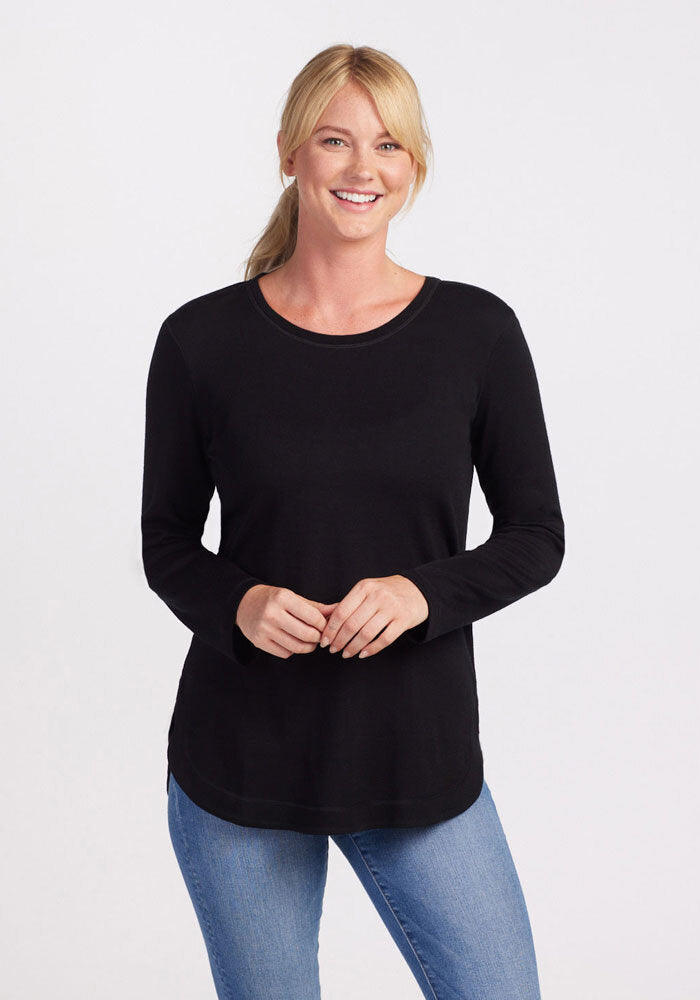 A woman with blonde hair tied back in a ponytail is smiling at the camera. She is wearing the Woolx Blair Tunic - Black, an ultra-cozy, long-sleeve top made from Australian Merino Wool, paired with light blue jeans. Her hands are clasped together in front of her against a plain white background. | Karly is 5'10', wearing a size S