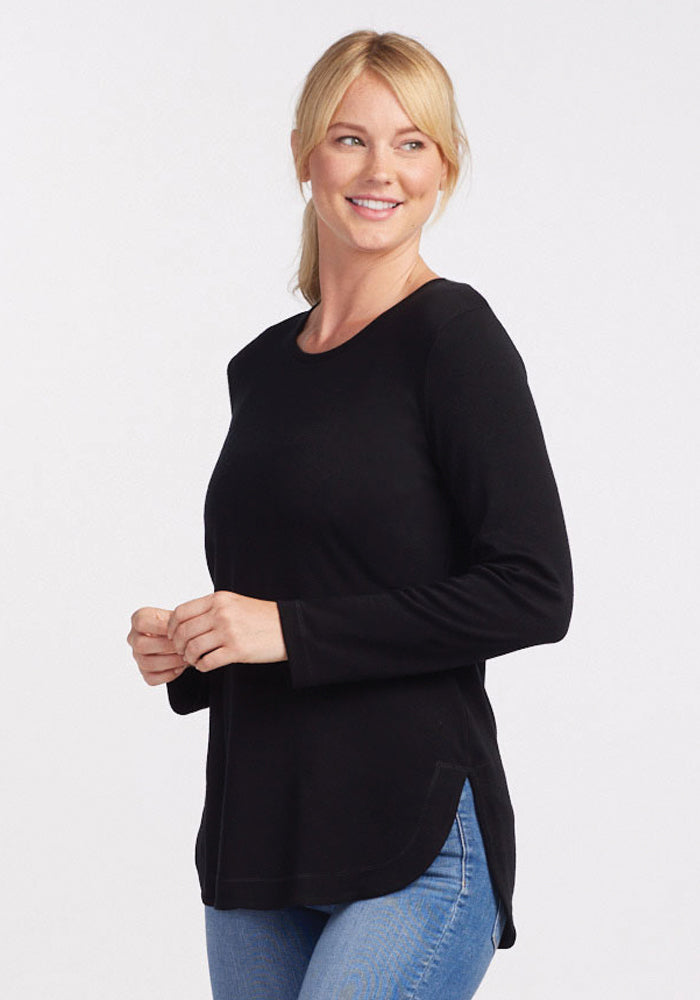 A person wearing the Woolx Blair Tunic in black and blue jeans smiles while looking to the right. The tunic features a rounded hem with side slits. The background is plain white.