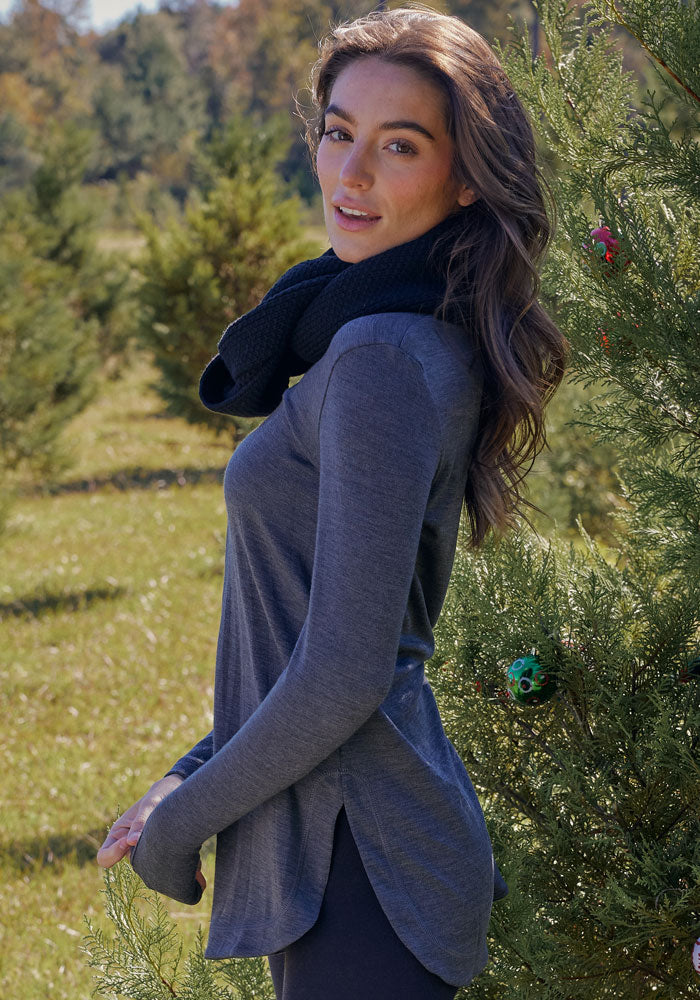 A woman with long brown hair stands by a pine tree, smiling over her shoulder in sunlight, wearing the Woolx Blair Tunic in Cinder Melange and a black scarf—ideal for travel.