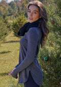 A woman with long brown hair stands by a pine tree, smiling over her shoulder in sunlight, wearing the Woolx Blair Tunic in Cinder Melange and a black scarf—ideal for travel.