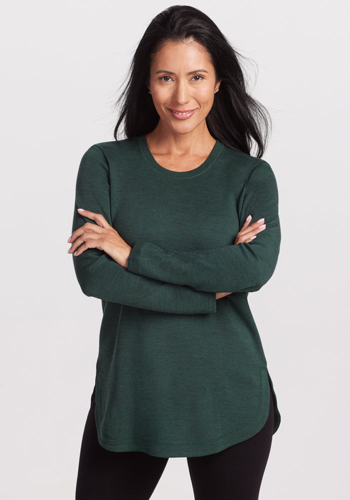 A woman with long dark hair wears the Woolx Blair Tunic • Final Sale - Forest and black pants, standing with arms crossed and smiling against a white background. This Merino wool tunic is odor-resistant and provides lightweight warmth for all-day comfort.