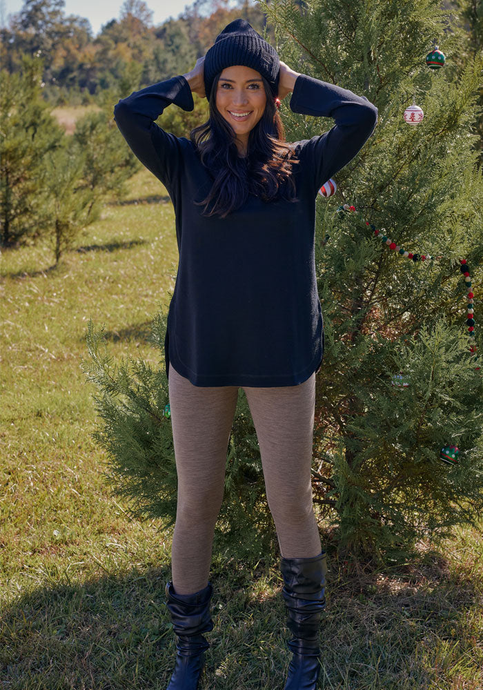 A woman stands outside in front of a decorated evergreen, smiling with her hands on her black knit hat. She wears the Woolx Blair Tunic in Black with tan leggings and black boots, surrounded by grass and trees.