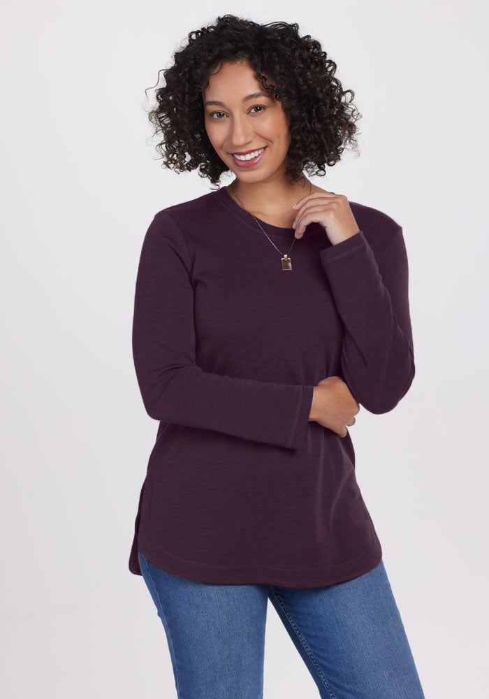 A smiling woman with curly hair models the Woolx Blair Tunic in Deep Plum—final sale—paired with blue jeans, standing against a white background, one hand near her chin and the other resting on her arm.