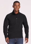 A man wears the Woolx Blizzard 1/4 Zip in black with blue jeans, standing against a light background and smiling gently with his hands relaxed at his sides. | Trell is 6'2", wearing a size M