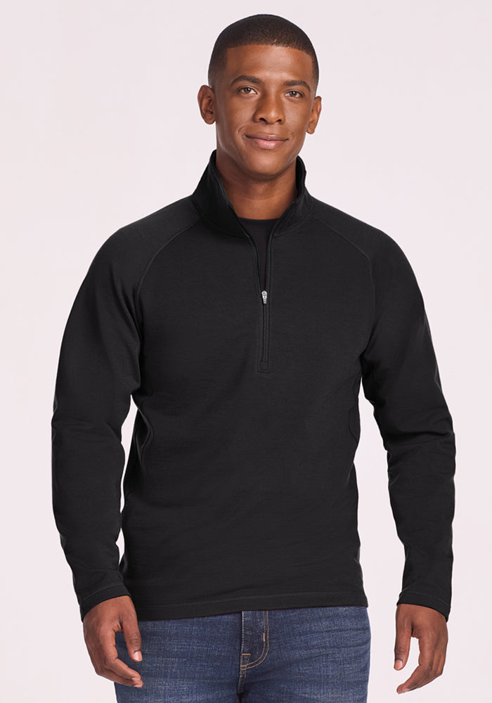 A man wears the Woolx Blizzard 1/4 Zip in black with blue jeans, standing against a light background and smiling gently with his hands relaxed at his sides. | Trell is 6'2", wearing a size M