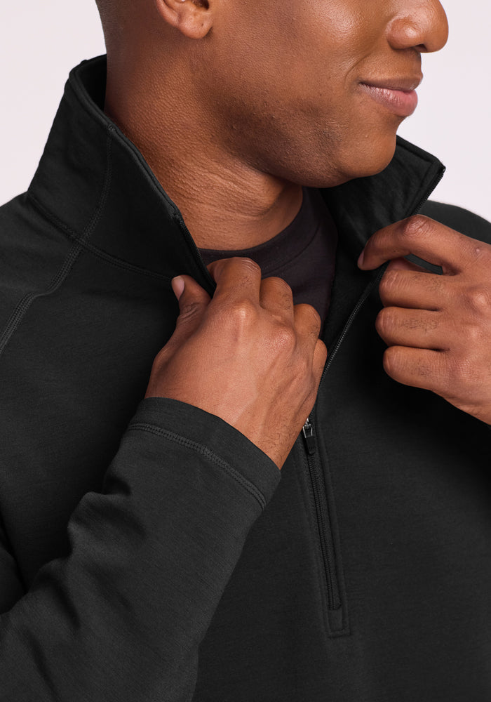 A person smiles slightly while adjusting the collar of the Woolx Blizzard 1/4 Zip in black, showing a black undershirt underneath.