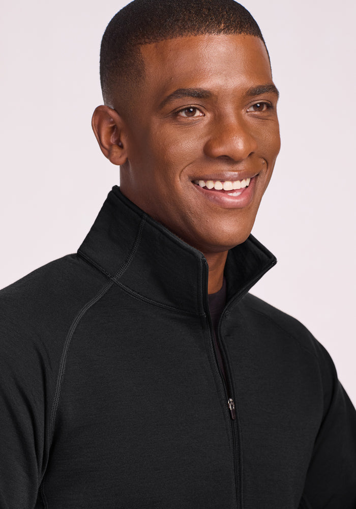 A smiling man with short hair wears the Woolx Blizzard 1/4 Zip in black, featuring a high collar, against a light background.