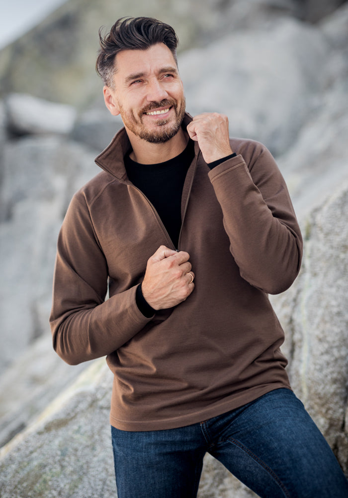 A man with short dark hair and a beard smiles while sitting on rocks outdoors. He wears a Woolx Blizzard 1/4 Zip - Black sweater over a black shirt and blue jeans, pulling up the collar of his cozy, moisture-wicking top.
