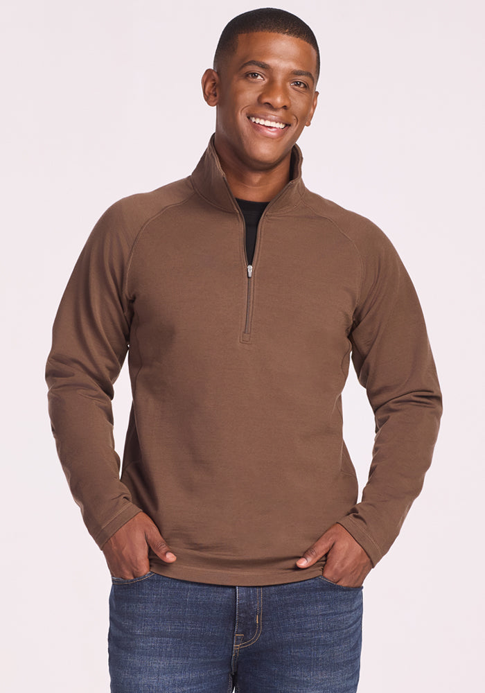 A smiling man wears the Woolx Blizzard 1/4 Zip in Bourbon Oak and blue jeans, standing with his hands in his pockets against a light background.
