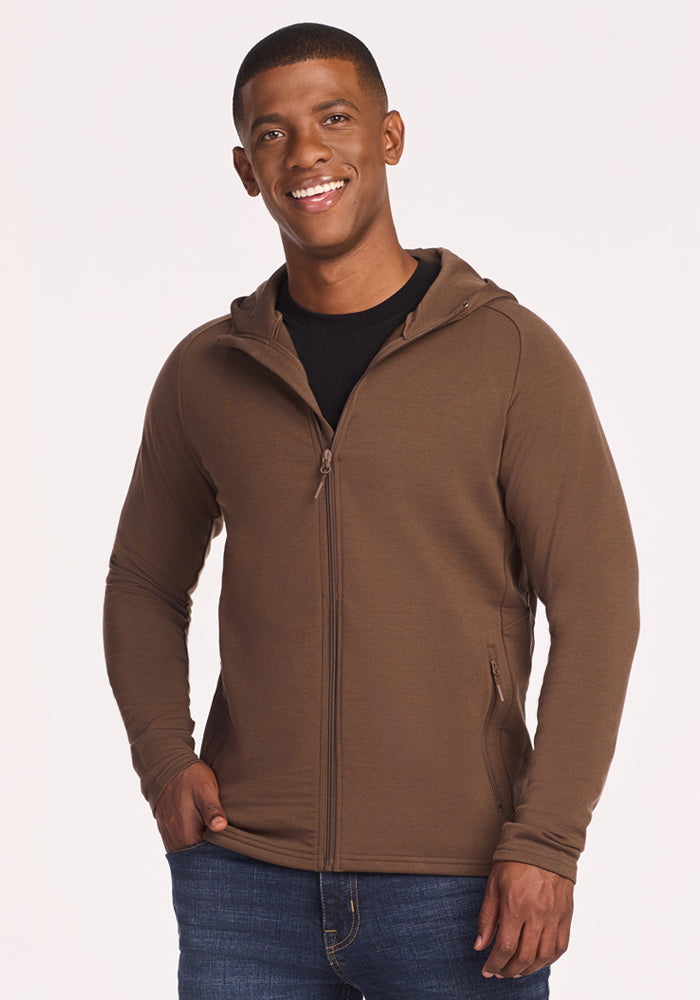 A smiling man wears the Woolx Boulder Hoodie • Final Sale - Bourbon Oak over a black shirt and blue jeans, standing with one hand in his pocket against a plain light background.