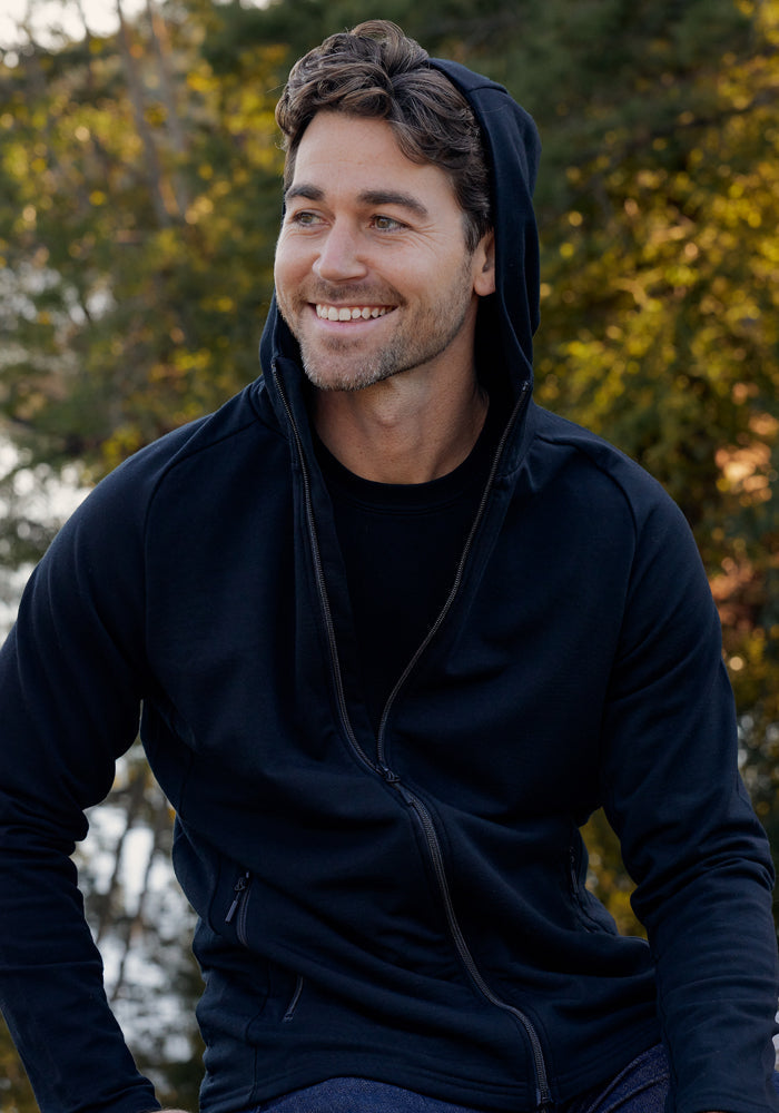 A smiling man with wavy brown hair wears the Woolx Boulder Hoodie in black, hood up, while sitting outdoors among green trees and sunlight.