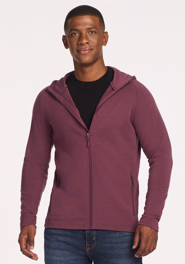 A man faces forward, smiling slightly, wearing the Woolx Boulder Hoodie in Grape Harvest over a black t-shirt and blue jeans against a plain light background.