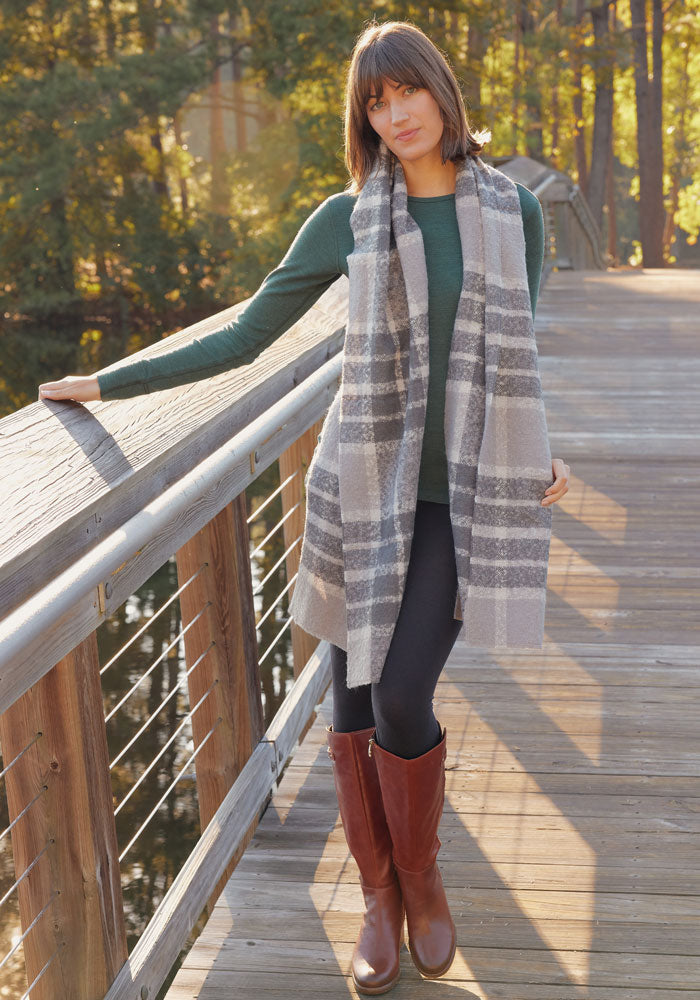 A woman in a green long-sleeve top, black leggings, brown boots, and the Woolx Bradie Scarf in Dark Grey Plaid leans on a wooden bridge railing in a sunlit forest.
