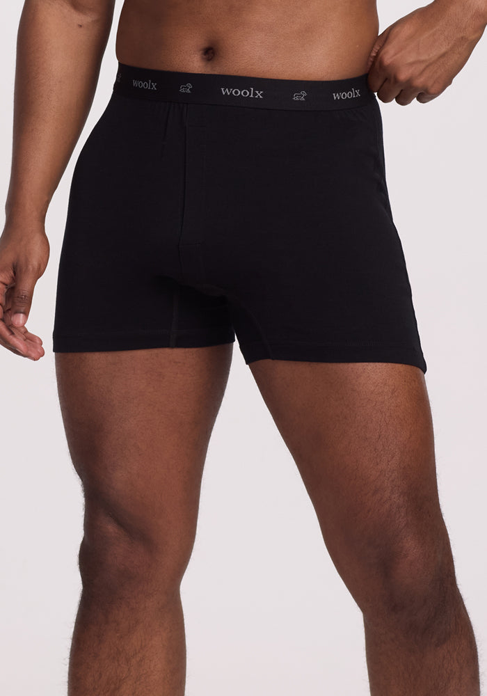 A person is wearing Breathe Boxer Shorts - Black from Woolx, featuring a "woolx" waistband, highlighting the breathability of Australian Merino Wool. The image shows them from mid-thigh to the waist against a plain background. | Trell is 6’2”, wearing a size M