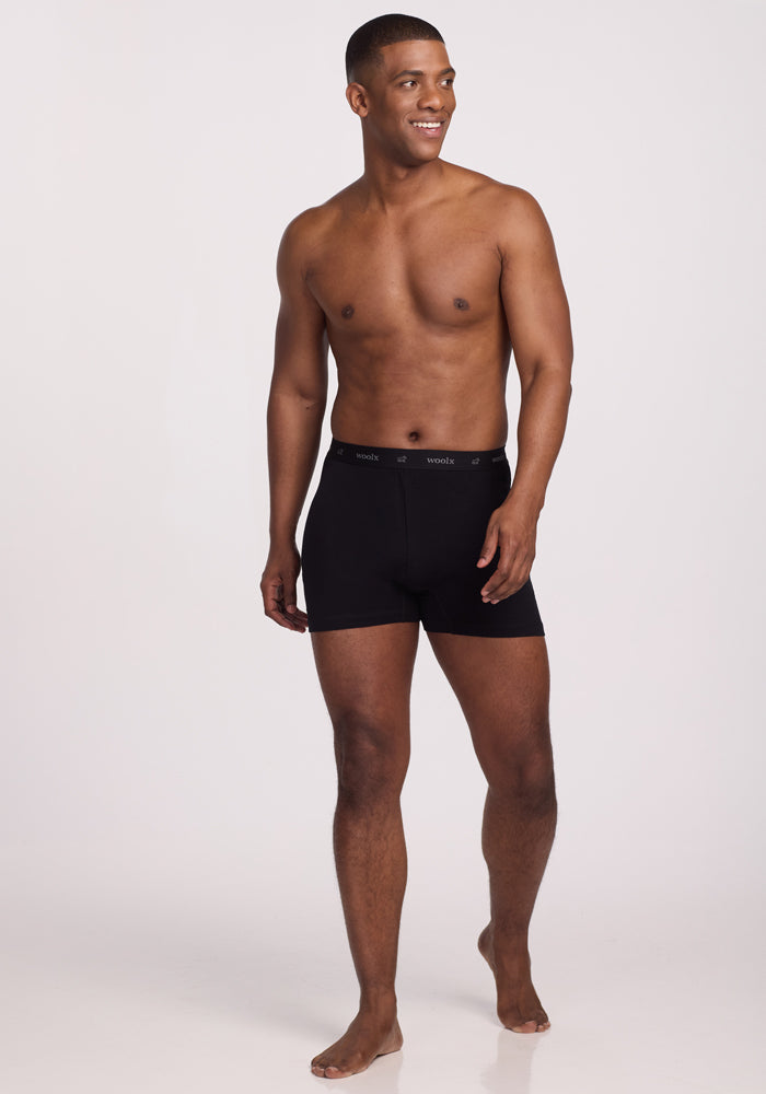 A man stands barefoot against a plain white background, smiling. He is wearing Woolx Breathe Boxer Shorts - Black, crafted from Australian Merino wool for optimal breathability, while being shirtless.