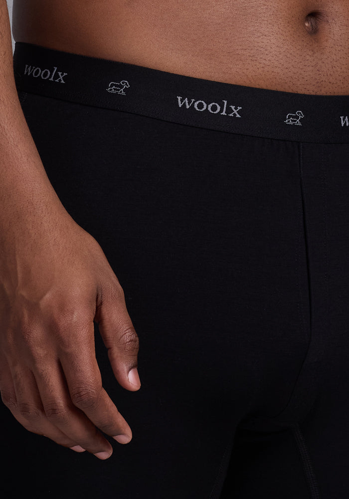 A close-up shows someone wearing Woolx Breathe Boxer Shorts in black. The breathable waistband with the logo and small sheep icons sits below their waist, while they rest a hand by their side against a neutral background. These Woolmark® Certified shorts provide premium comfort.
