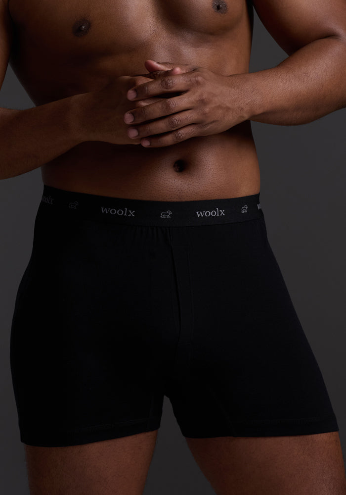 Against a dark backdrop, a person models the Breathe Boxer Shorts - Black by Woolx, featuring a Woolmark® Certified waistband with "woolx" inscribed. Their hands are clasped in front.