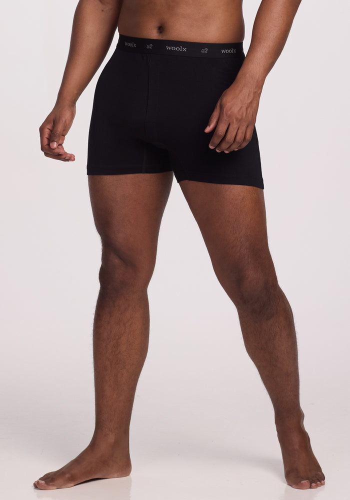 A person stands barefoot against a white background wearing Woolx Breathe Boxer Shorts - Black. The breathable lightweight Merino Wool and texted waistband are highlighted, with their relaxed posture emphasizing comfort in a neck-down capture.
