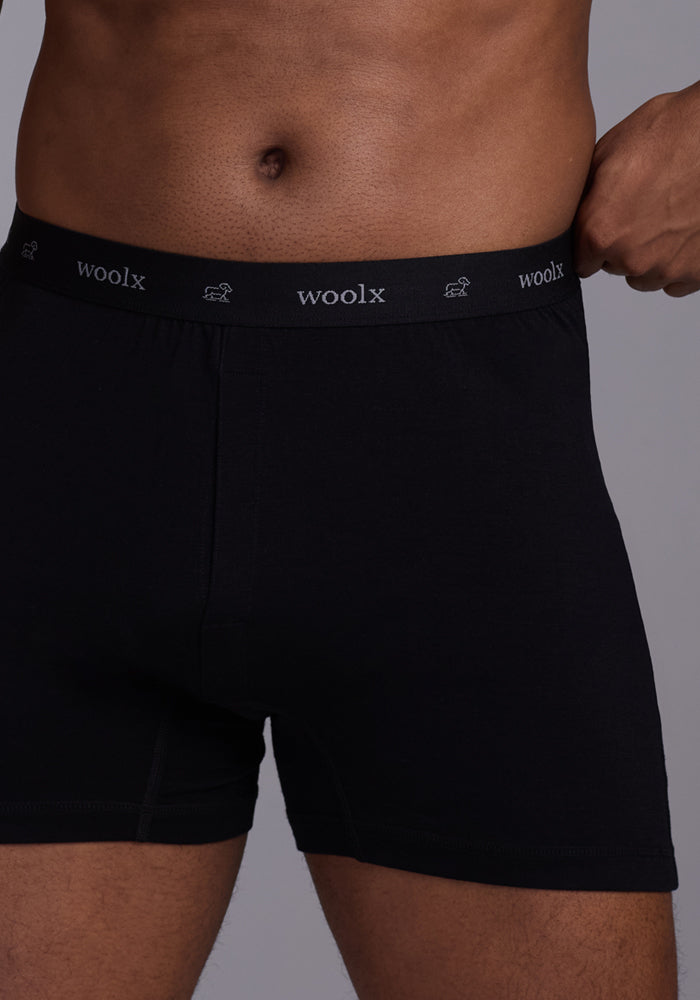 A person wearing Woolx's Breathe Boxer Shorts - Black, featuring a "Woolx" branded waistband with small sheep icons and Woolmark® Certification, stands against a gray background. The image is cropped to show from torso to mid-thigh.