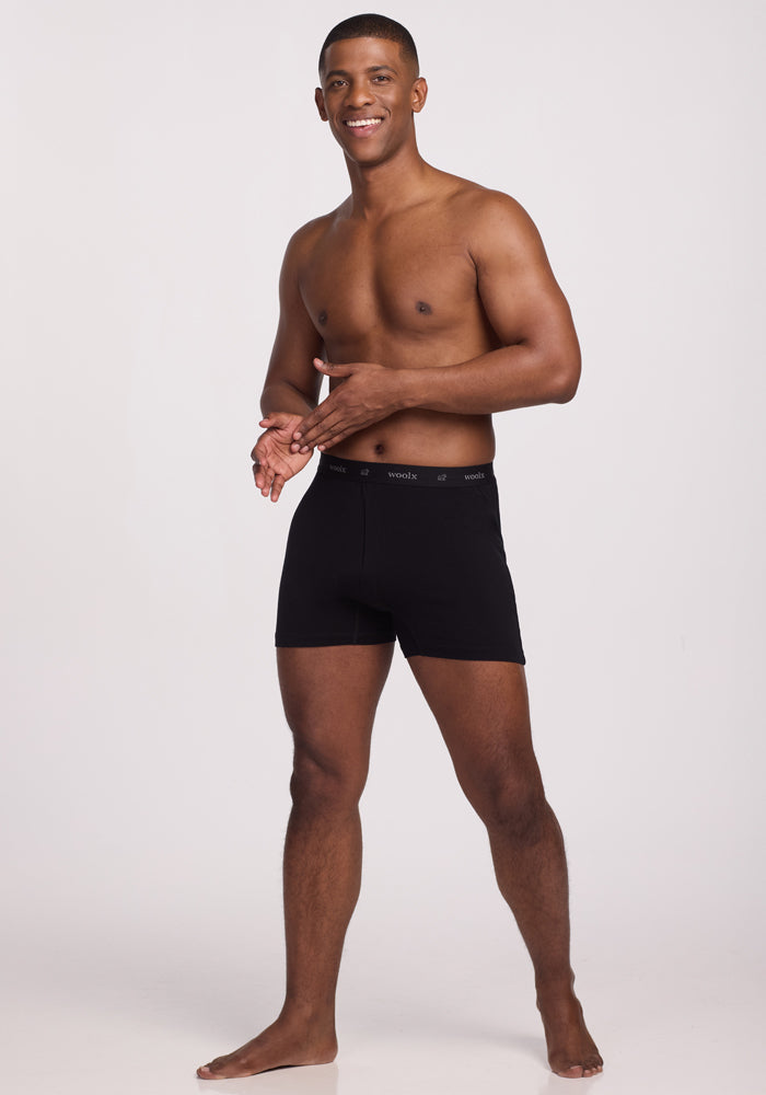 A smiling man stands barefoot against a plain background, wearing Woolx's Breathe Boxer Shorts in Black. His hands appear to be adjusting or showcasing the waistband.