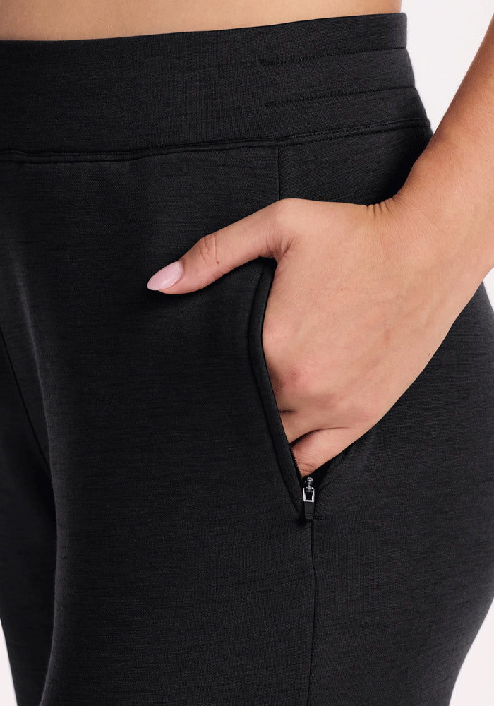 A close-up shows a hand with light pink nail polish resting in the front zippered pocket of Woolx Bree Lounge Pants - Black, highlighting the cozy fabric and waistband against a plain background.