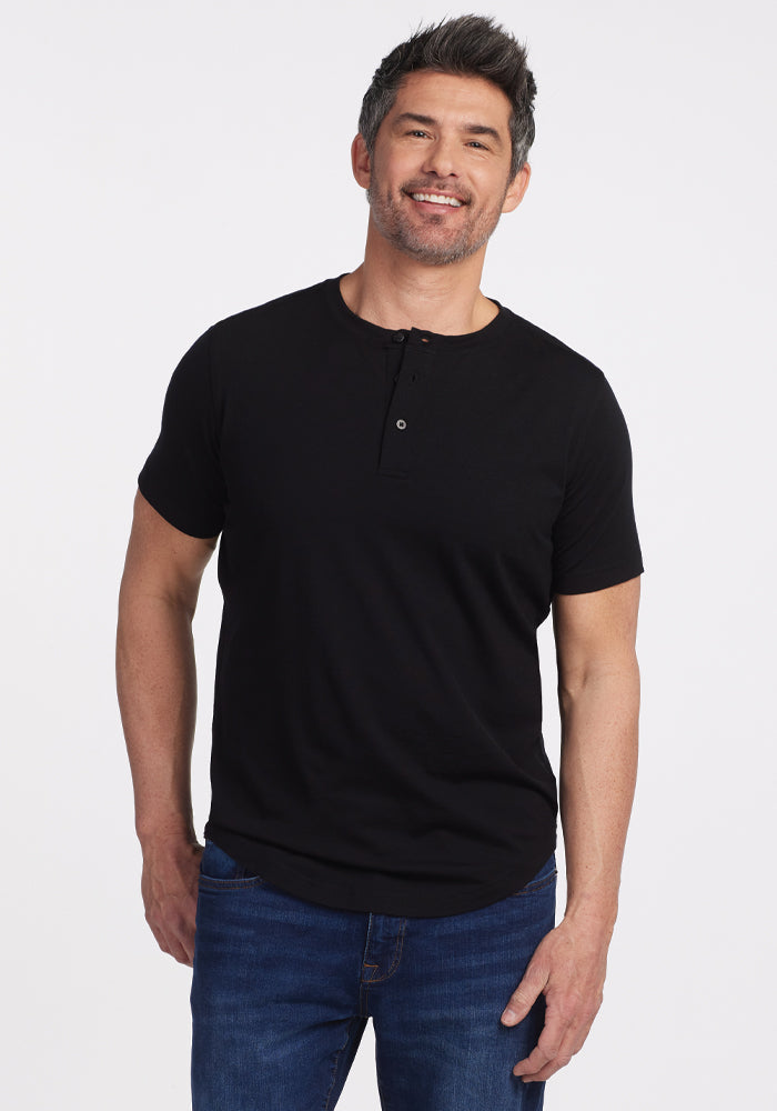 A smiling man with short, gray-streaked hair wears a black Bronx Henley from Woolx and blue jeans. He stands against a plain white background with one hand in his pocket, exuding casual elegance. | Matthew is 6’, wearing a size L