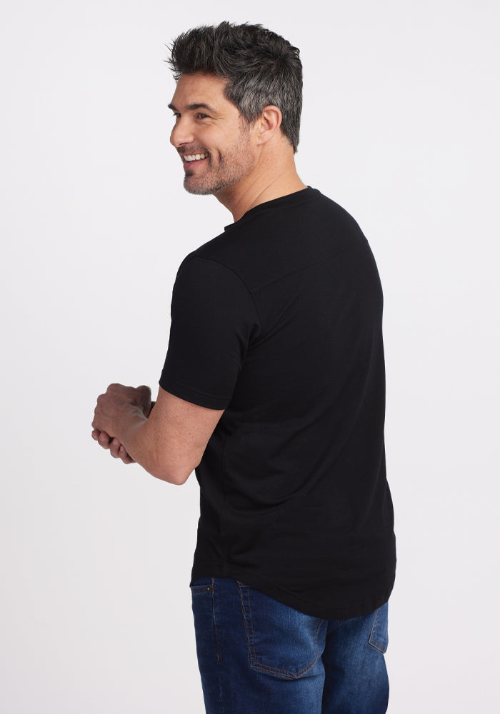 A smiling man with gray hair, wearing a Bronx Henley - Black from Woolx, crafted from Woolmark Certified Merino wool, along with blue jeans, is turned slightly to his right. He looks over his shoulder at the camera against a plain white background.