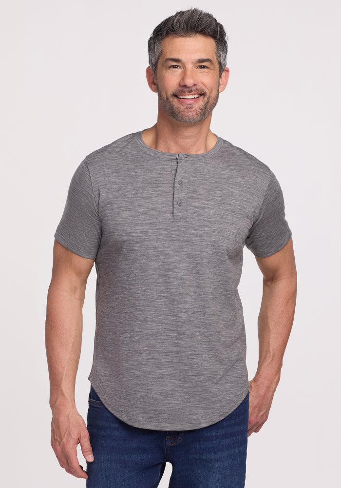 A smiling man with short gray hair and a trimmed beard wears the Woolx Bronx Henley • Final Sale - Graphite Heather and blue jeans. He stands against a plain light background with one hand relaxed by his side.