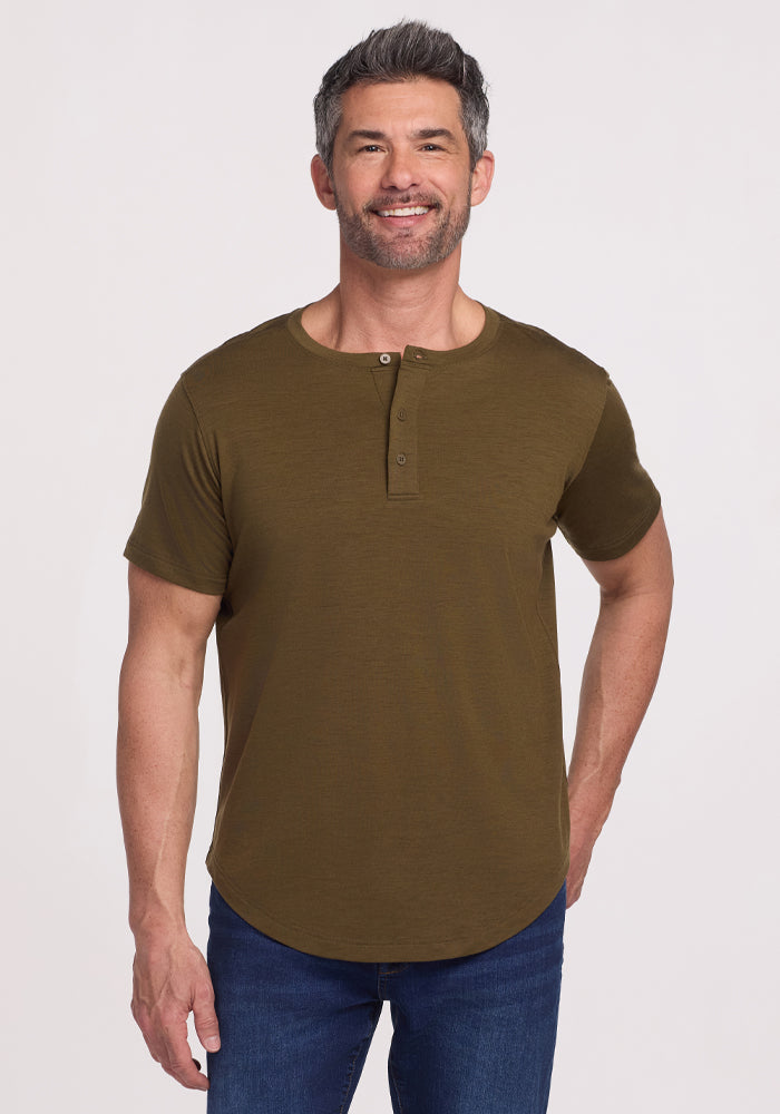 A man with short dark hair and a beard smiles while wearing the Woolx Bronx Henley • Final Sale - Olive Leaf and blue jeans, standing against a plain white background.