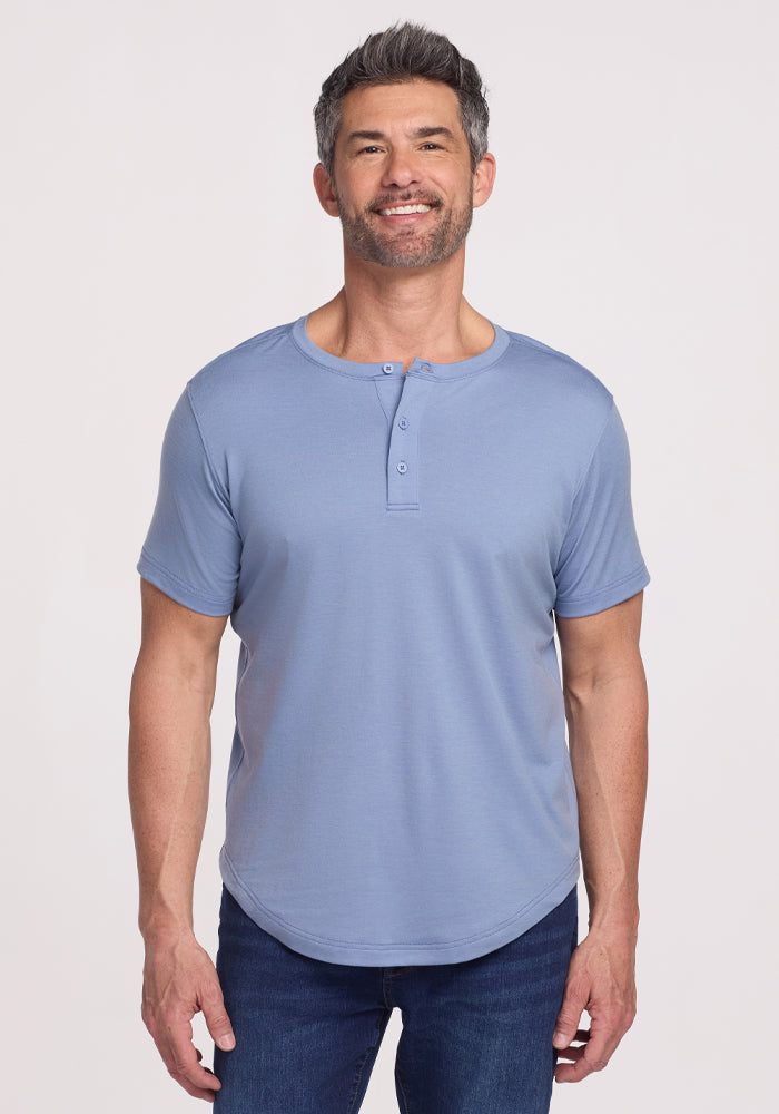 A man with short dark hair and a beard smiles while wearing the Woolx Bronx Henley • Final Sale - Stonewash, paired with dark jeans, standing against a plain light background.