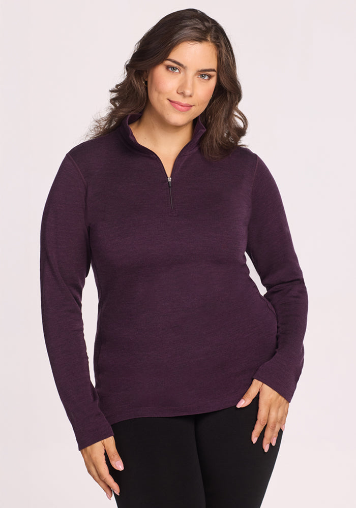 A woman with wavy brown hair stands against a plain light background, smiling softly with one hand on her hip, wearing the Woolx Brooke 1/4 Zip • Final Sale - Deep Plum and black pants.