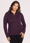 A woman with wavy brown hair stands against a plain light background, smiling softly with one hand on her hip, wearing the Woolx Brooke 1/4 Zip • Final Sale - Deep Plum and black pants.