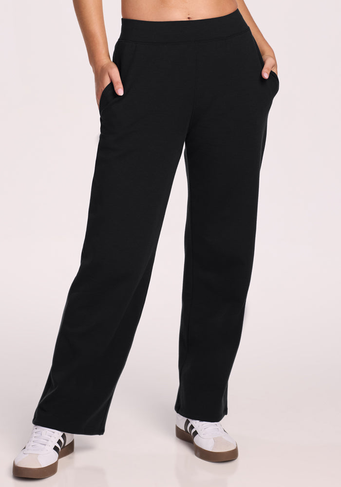 A person wears Woolx Brynn Pants • Final Sale - Black, styled with hands in pockets and white sneakers with brown soles, standing against a light background.