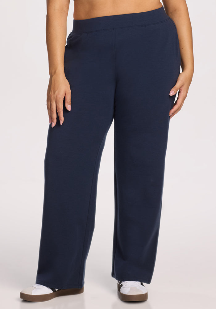 A person stands against a plain light background, wearing Woolx Brynn Pants • Final Sale - Deep Navy and white sneakers. The image is cropped from the waist down.