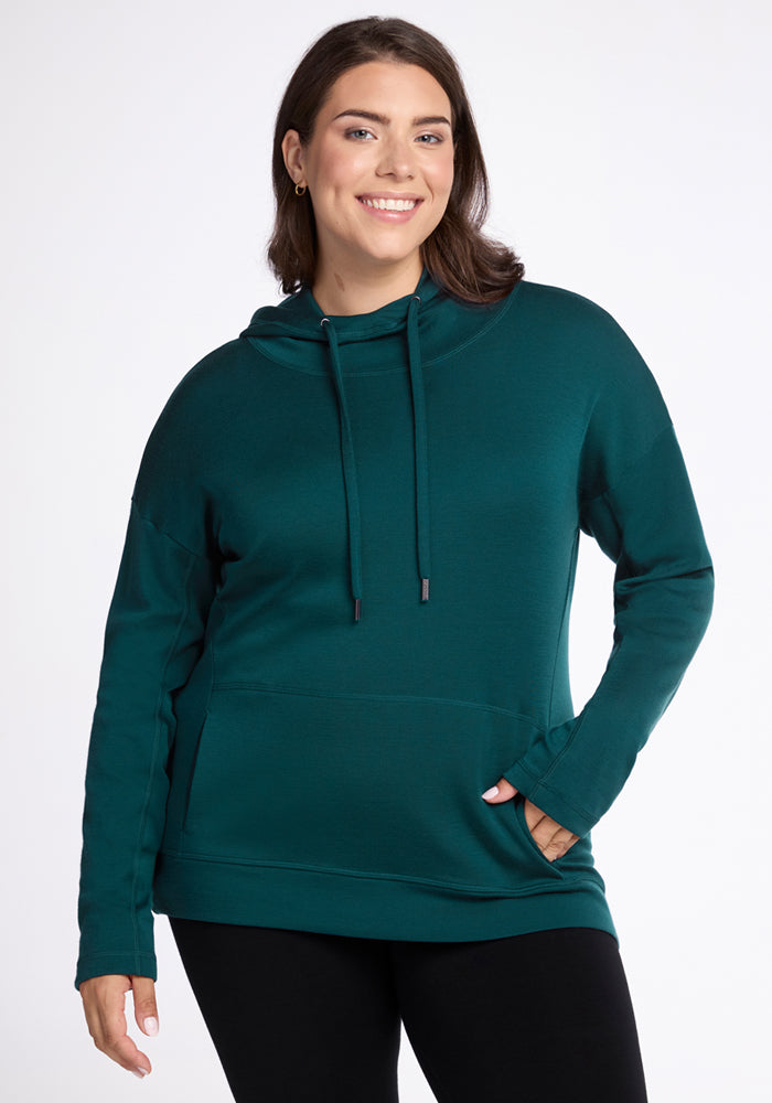 A woman with long brown hair smiles while wearing the Woolx Callie Hoodie in Riverbank Green, an ultra-soft hooded sweatshirt with a front pocket, and black leggings, standing against a plain white background.