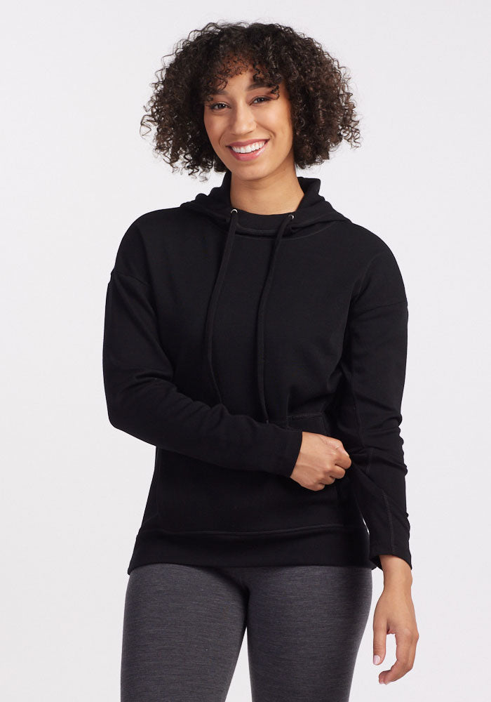 A person with curly hair is smiling while wearing a cozy Callie Hoodie - Black from Woolx and gray leggings. They are standing against a plain white background with one hand touching their hip and the other resting on their stomach. | Tori is 5'7", wearing a size S