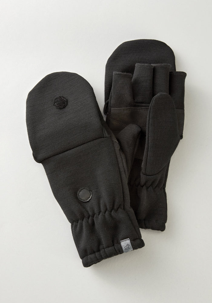 The Cameron Glove Mitten in black from Woolx is displayed on a light surface. Made with a Merino wool blend, these mittens provide exceptional warmth. They feature visible finger slots under the mitten cover, which can be folded back and secured with Velcro. Ribbed cuffs and a small logo tag offer subtle detailing.