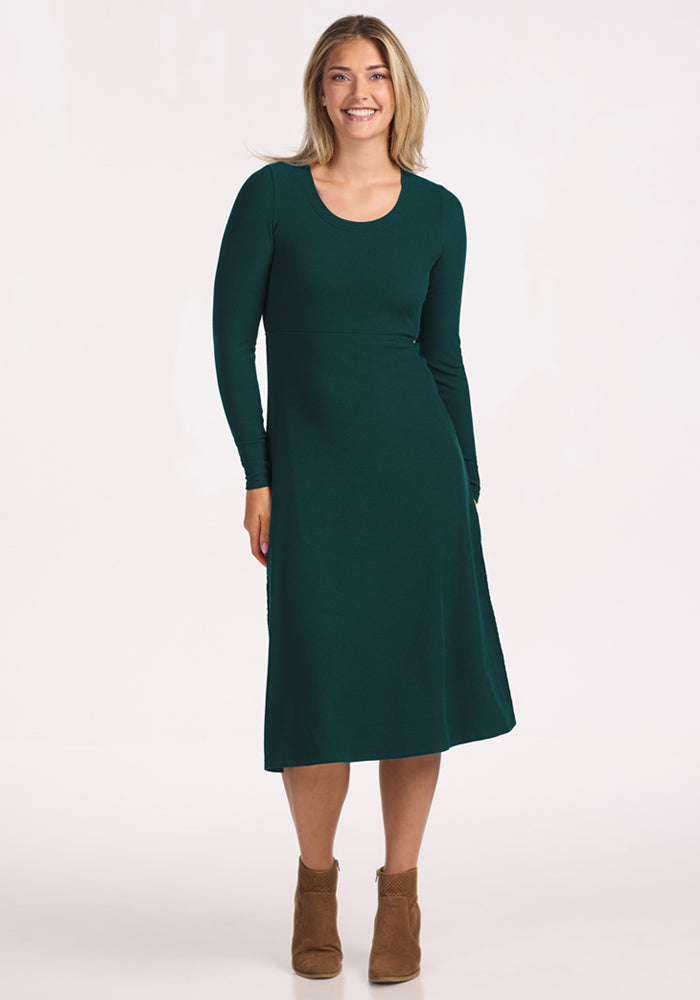 A woman smiles against a light background, wearing the Woolx Caroline Ribbed Twirl Dress in Deep Timber with brown ankle boots. | Ariana is 5'11", wearing a size S