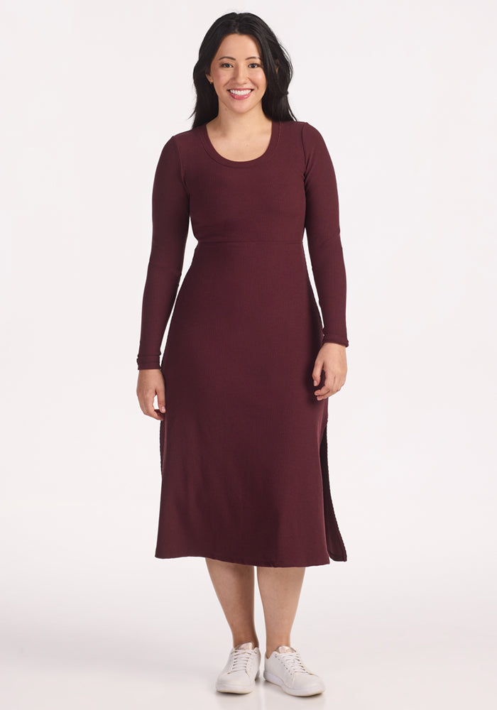 A woman with long dark hair, wearing the Woolx Caroline Ribbed Twirl Dress in Toasted Garnet and white sneakers, smiles against a plain white background. | Sarah is 5'8", wearing a size S
