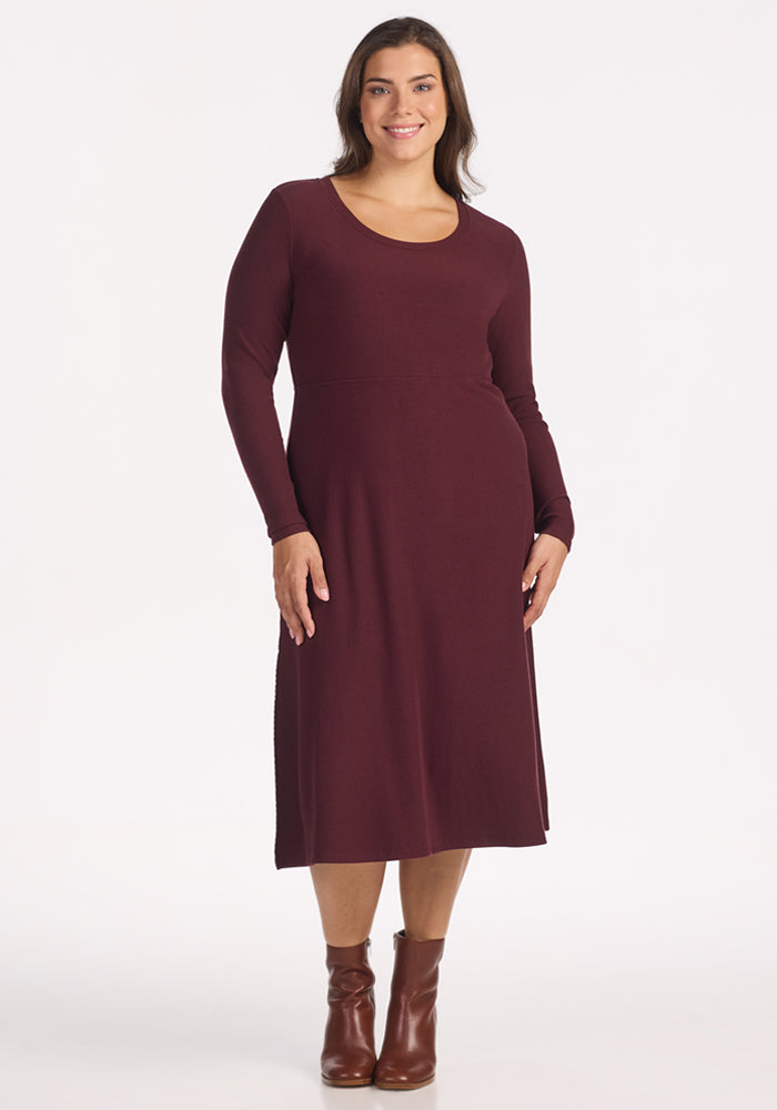 A woman with long brown hair models the Woolx Caroline Ribbed Twirl Dress in Toasted Garnet, featuring a wrinkle-resistant, versatile silhouette and paired with brown ankle boots, smiling against a plain white background. | Juliana is 5'10", wearing a size XL